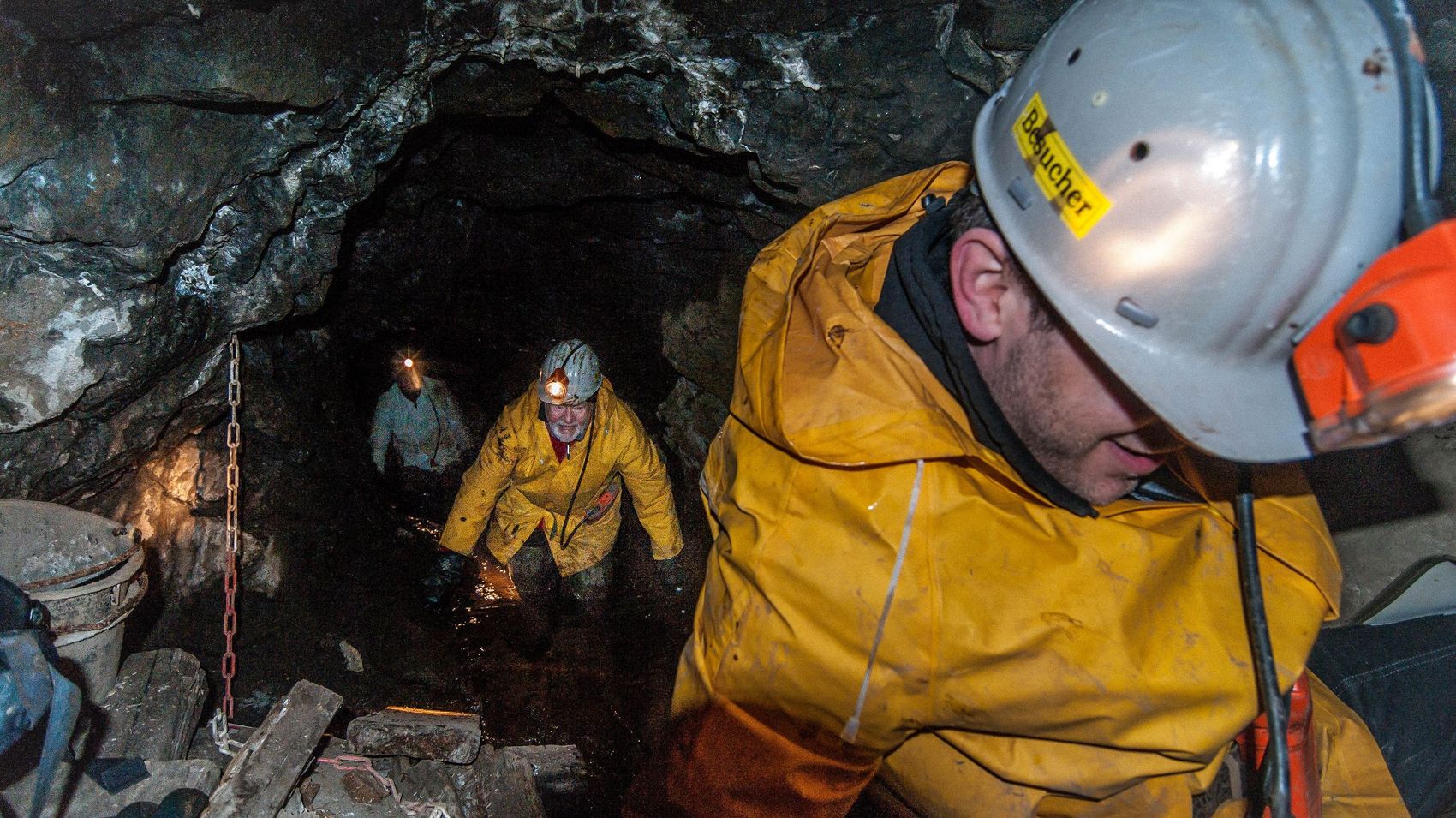 Ausschnitt unter Tage Drei Bergarbeiter sind mit Grubenhelm und Grubenlampe unter Tage im Stollen unterwegs.