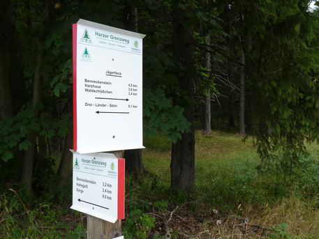 Wegeschild Ein weißes Wegeschild mit roter Umrandung zum Grenzweg im Wald. Hinter dem Schild sind Bäume zu sehen.