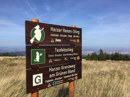 Ein Informationsschild über Länge, Ausschilderung und Start/Endpunkt der Themenwanderwege Harzer Hexenstieg, Harzer Grenzweg und Teufelsstieg, auf einer Wiese. Im Hintergrund hat man Aussicht über die Landschaft.