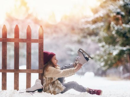 Ein kleines Mädchen sitzt auf schneebedecktem Boden angelehnt an einen Zaun und zieht sich Schlittschuhe an. Im hintergrund scheint die Sonne durch die Bäume.